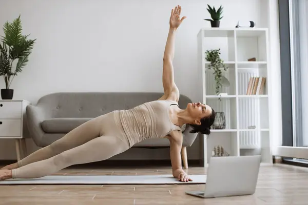 Mature woman in tight sports clothes taking up Side Plank Pose on living room floor with rubber mats during yoga practice. Active slim lady building overall flexibility using Vasisthasana exercise.
