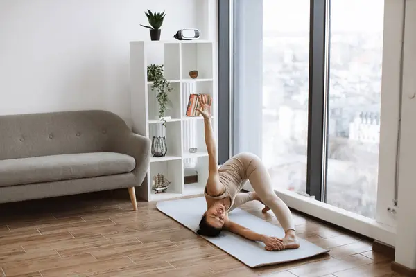 Side view of stunning mature woman wearing beige sportswear lying on mat and breeds legs widely. Slim caucasian brunette practicing yoga pose at home with panoramic windows.