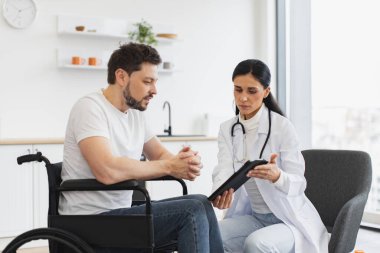 Hemşire, tekerlekli sandalyedeki olgun bir adamla konuşuyor. Modern tablet kullanarak sınav sonuçlarını gösteriyor. Esmer beyaz kadın doktorun portresi evde sakallı bir erkek hastanın durumunu kontrol ediyor..