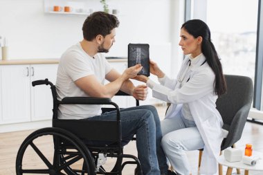 Yetişkin beyaz kadın doktor evde sakallı bir erkek hasta var mı diye bakıyor. Genç bayan hemşire olgun tekerlekli sandalye kullanıcısıyla konuşuyor, modern tablette omurga röntgeni gösteriyor..
