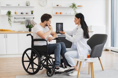 Beyaz kadın doktor portresi evde sakallı bir erkek hastanın durumunu kontrol ediyor. Hemşire olgun tekerlekli sandalye kullanıcısıyla konuşuyor, modern tablette omurga röntgeni gösteriyor..