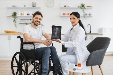 Genç kadın hemşire ve olgun tekerlekli sandalye kullanıcısı portresi modern tablette omurga röntgeni gösteriyor. Kafkasyalı kadın doktor evde sakallı bir erkek hastanın durumunu kontrol ediyor..