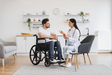 Kadın doktor, uzman kontrolleri yapıyor ve evde, mutfakta özürlü bir hastaya ilaç veriyor. Tıbbi geçmişle ilgili tavsiyeleri dinleyen odaklı tekerlekli sandalye kullanıcısı.