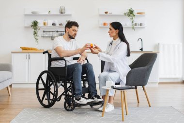 Kadın doktor, uzman kontrolleri yapıyor ve evde, mutfakta özürlü bir hastaya ilaç veriyor. Tıbbi geçmişle ilgili tavsiyeleri dinleyen odaklı tekerlekli sandalye kullanıcısı.