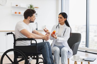 Tıbbi geçmişle ilgili tavsiyeleri dinleyen odaklanmış tekerlekli sandalye kullanıcısı. Kadın doktor, evde, mutfakta engelli bir hastaya uzman muayenesi ve ilaç veriyor..