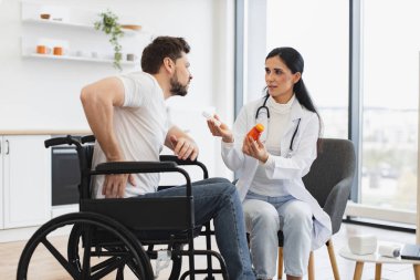 Engelli hastayla iletişim kuran kadın pratisyen hekimin elinde ilaç şişesi var. Doktor, omurgasının alt kısmında ağrısı olan beyaz bir erkekle sağlık sorunlarını tartışıyor..