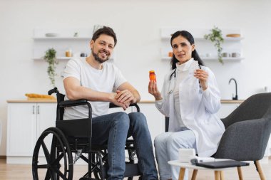 Beyaz üniformalı kadın terapistin portresi. Elinde haplar var. Evde, açık mutfakta, tekerlekli sandalyedeki engelli bir hastaya danışıyor..