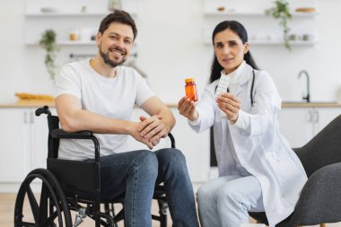 Beyaz üniformalı kadın terapistin portresi. Elinde haplar var. Evde, açık mutfakta, tekerlekli sandalyedeki engelli bir hastaya danışıyor..