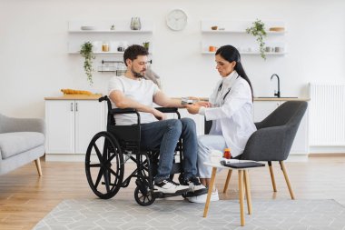 Çekici kardiyolog engelli bir hastaya evde hipertansiyon olmasının tüm nedenlerini anlatıyor. Tekerlekli sandalyedeki yetişkin adam sağlıklı bir hayat sürmenin gerekliliğini anlatıyor..