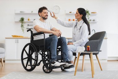 Kendine güvenen 30 yaşındaki kadın doktor ev ziyareti sırasında kızıl ötesi alın termometresi kullanarak tekerlekli sandalyedeki olgun sakallı hastanın vücut ısısını ölçüyor..