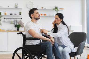 Pratisyen doktor evde düzenli kontrol yaparken kalp atışlarını inceliyor. Beyaz önlüklü kadın steteskop kullanırken, yetişkin adam tekerlekli sandalyede oturuyor..