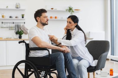 Pratisyen doktor evde düzenli kontrol yaparken kalp atışlarını inceliyor. Beyaz önlüklü kadın steteskop kullanırken, yetişkin adam tekerlekli sandalyede oturuyor..