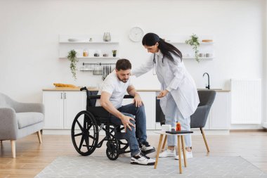 Doktor önlüklü kadın tekerlekli sandalyeden çıkan sakallı adamı destekliyor. Evde fizyoterapi gördüğü için sırtından yaralanan bir sağlık uzmanı..