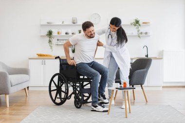 Evde fizyoterapi gördüğü için sırtından yaralanan bir sağlık uzmanı. Doktor önlüklü kadın. Tekerlekli sandalyeden çıkan sakallı adamı destekliyor..