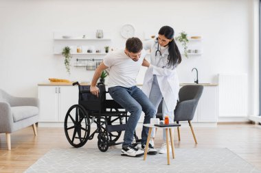 Fizikçi ona modern bir evde el uzatırken, tekerlekli sandalyenin yanında duran adamın tam boy görüntüsü. Başarılı bir tedaviden sonra hastayı engelli olarak teşvik eden tıbbi kadın..