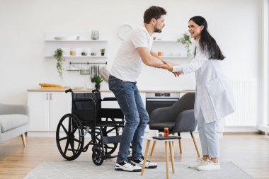 Başarılı bir tedaviden sonra hastayı engelli olmaya teşvik ediyor. Tekerlekli sandalyenin yanında duran adamın geniş açılı görüntüsü. Doktorlar ona modern evde el uzatıyor..