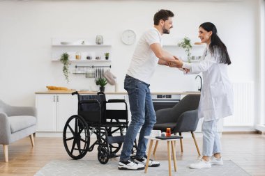 Beyaz tenli çekici bir kadının, engelli genç bir hastaya modern aydınlık mutfakta tekerlekli sandalyeden kalkması için yardım etmesi. Evdeki oturma odasında hasta destekleyen bir tıp uzmanı..