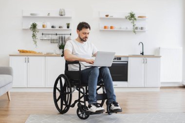 Tekerlekli sandalyedeki neşeli beyaz adam dizlerinin üzerinde dizüstü bilgisayar tutarken açık planlı mutfağın ortasında kalıyor. Evde geçirdiği kazadan sonra iyileşme sürecinde internet sayfalarında mutlu genç sörfçüler..