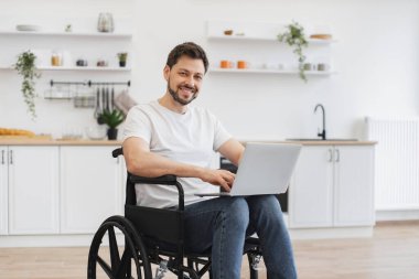 Tekerlekli sandalyedeki beyaz adamın portresi dizlerinin üzerinde dizüstü bilgisayar tutarken açık-plan mutfağın ortasında duruyor. Evde geçirdiği kazadan sonra iyileşme sürecinde internet sayfalarında mutlu genç sörfçüler.