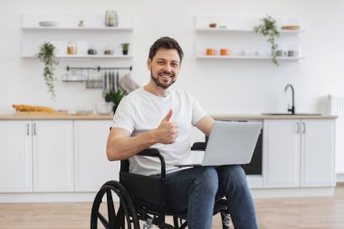 Kafkasyalı tekerlekli sandalyede dizlerinin üstünde dizüstü bilgisayar tutan beyaz adamın portresi. Parlak mutfakta başparmağını gösteriyor. Evde geçirdiği kazadan sonra iyileşme sürecinde internet sayfalarında mutlu genç sörfçüler.