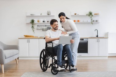 Tekerlekli sandalyedeki beyaz bir adam dizlerinin üzerinde dizlerinin üzerinde karısının çalışma sonuçlarını gösteriyor. Evde geçirdiği kazadan sonra iyileşme sürecinde internet sayfalarında mutlu sörfler..