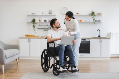 Tekerlekli sandalyedeki beyaz adam dizlerinin üzerinde diz çöküp karısı için parlak mutfağın ortasında iş sonuçlarını gösteriyor. Evde geçirdiği kazadan sonra iyileşme sürecinde internet sayfalarında mutlu sörfler..