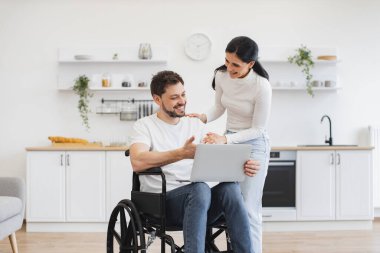 Evde geçirdiğin kazadan sonra internet sitelerinde mutlu sörfler. Tekerlekli sandalyedeki beyaz adam dizlerinin üzerinde dizlerinin üzerinde karısı için parlak mutfağın ortasında iş sonuçlarını gösteriyor..