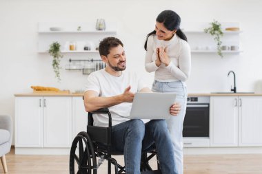 Evde geçirdiğin kazadan sonra internet sitelerinde mutlu sörfler. Tekerlekli sandalyedeki beyaz adam dizlerinin üzerinde dizlerinin üzerinde karısı için parlak mutfağın ortasında iş sonuçlarını gösteriyor..