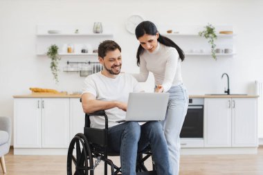 Evde geçirdiğin kazadan sonra internet sitelerinde mutlu sörfler. Tekerlekli sandalyedeki beyaz adam dizlerinin üzerinde dizlerinin üzerinde karısı için parlak mutfağın ortasında iş sonuçlarını gösteriyor..