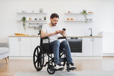 Modern apartman mutfağında dinlenirken akıllı telefon kullanan, engelli beyaz bir erkek. Günlük giysiler içindeki neşeli kişi evdeki dijital cihazdan mesaj gönderiyor..