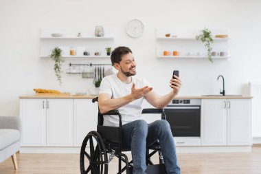 Modern apartmanda gezmeyi planlarken engelli cep telefonlu neşeli genç bir yetişkin. Gülümseyen beyaz adam yemek odasında video başvurusu aracılığıyla akrabalarıyla sohbet ediyor..
