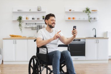 Akrabalarıyla sohbet eden gülümseyen beyaz bir adam yemek odasında video çağrı uygulaması aracılığıyla. Modern apartmanda gezmeyi planlarken engelli cep telefonlu neşeli genç bir yetişkin..