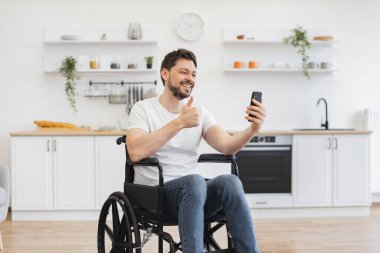 Akrabalarıyla sohbet eden gülümseyen beyaz bir adam yemek odasında video çağrı uygulaması aracılığıyla. Modern apartmanda gezmeyi planlarken engelli cep telefonlu neşeli genç bir yetişkin..