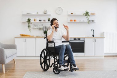 Modern stüdyo dairesinin rahatlığını kullanırken tekerlekli sandalyedeki yetişkin birinin cep telefonuyla konuşmasının geniş açılı görüntüsü. Olgun Kafkasyalı adam hastalıktan kurtulurken doğum günü partisi düzenliyor..