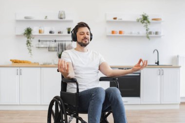 Rahatlamış yetişkin ve engelli. Meditasyon yoluyla sağlıklı yaşam tarzını destekliyor. Mutfak arka planında nilüfer pozisyonunda tekerlekli sandalyede dinlenen, kulaklı, gülümseyen beyaz adamın portresi..