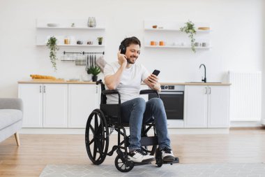 Beyaz tişörtlü neşeli adam stüdyo dairesindeki tekerlekli sandalyede oturan akıllı telefona bakarken elini kaldırıyor. Neşeli Kafkasyalı yetişkin kulaklıkla yüksek sesli melodiler dinlerken dans ediyor..