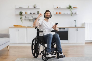 Mutfakta dijital aletlerle otururken tekerlekli sandalyedeki heyecanlı genç adam elleri havada melodiden havaya hareket ettiriyor. Kablosuz kulaklıklarla zengin ses deneyiminden zevk alan beyaz bir yetişkin..