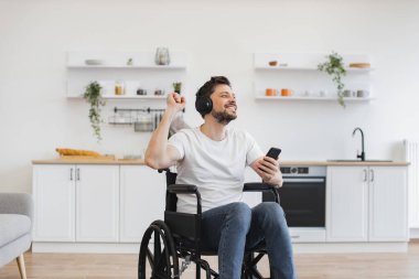 Mutfakta dijital aletlerle otururken tekerlekli sandalyedeki heyecanlı genç adam elleri havada melodiden havaya hareket ettiriyor. Kablosuz kulaklıklarla zengin ses deneyiminden zevk alan beyaz bir yetişkin..