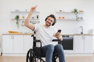 Kablosuz kulaklıklarla zengin ses deneyiminden zevk alan beyaz bir yetişkin. Mutfakta dijital aletlerle otururken tekerlekli sandalyedeki heyecanlı genç adam ellerini havaya kaldırıyor..
