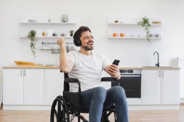 Kablosuz kulaklıklarla zengin ses deneyiminden zevk alan beyaz bir yetişkin. Mutfakta dijital aletlerle otururken tekerlekli sandalyedeki heyecanlı genç adam ellerini havaya kaldırıyor..