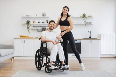 Kadın fizyoterapist ya da eş, tekerlekli sandalyedeki genç adamın modern dairelerde egzersiz yapmasına yardım ediyor. Üst düzey erkek hasta, hafif evde hastalık ve yaralanma sonrası rehabilitasyona giriyor..