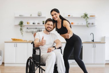 Kadın fizyoterapist ya da eş, tekerlekli sandalyedeki genç bir adama yardım ediyor. Dairelerde egzersizler yapıyor ve onaylıyor. Üst düzey erkek hasta, hafif evde hastalık ve yaralanma sonrası rehabilitasyona giriyor..