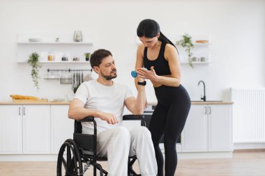 Engelli insanların rehabilitasyonu. Spor giyim sektöründe genç fizyoterapist ya da eş evde tekerlekli sandalye egzersizi yapan beyaz bir erkek hastaya yardım ediyor. Engelli, olgun, halterli adam eğitimi..