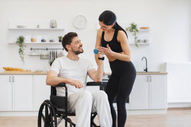 Spor giyim sektöründe genç fizyoterapist ya da eş evde tekerlekli sandalye egzersizi yapan beyaz bir erkek hastaya yardım ediyor. Engelli insanların rehabilitasyonu. Engelli, olgun, halterli adam eğitimi..