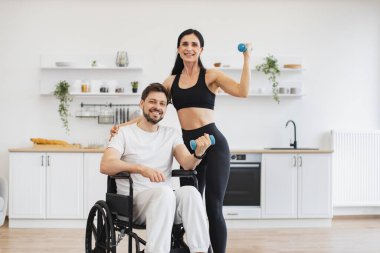 Engelli, olgun bir adamın karısıyla ya da kadın eğitmeniyle dambıl kullanırken çekilmiş portresi. Spor giyim sektöründe genç fizyoterapist ya da eş evde tekerlekli sandalye egzersizi yapan beyaz erkek hastaya yardım ediyor..