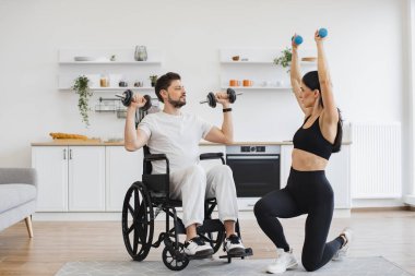 Spor giyim sektöründe genç fizyoterapist ya da eş evde tekerlekli sandalye egzersizi yapan beyaz bir erkek hastaya yardım ediyor. Engelli insanların rehabilitasyonu. Engelli, olgun, halterli adam eğitimi..