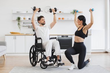 Spor giyim sektöründe milenyum fizyoterapisti tekerlekli sandalyedeki engelli bir adamın halterlerle antrenman yapmasına ve evdeki sakatlığından kurtulmasına yardım ediyor. Genç erkek hasta için rehabilitasyon.