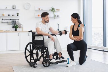 Spor kıyafetli bir fizyoterapist, tekerlekli sandalyedeki yetişkin bir adama evde halterlerle nasıl egzersiz yapıldığını gösteriyor. Erkek hasta, modern apartmanın arka planında hastalık ve yaralanma üzerine rehabilitasyon görüyor.