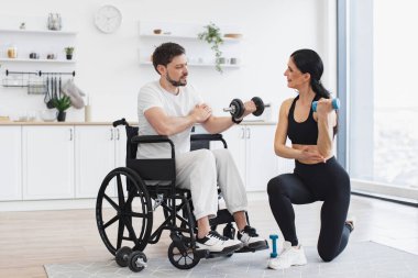 Erkek hasta, modern bir apartmanın arka planında hastalık ve yaralanma üzerine rehabilitasyona giriyor. Spor salonundaki fizyoterapist, tekerlekli sandalyedeki yetişkin bir adama evde dambıllarla nasıl egzersiz yapıldığını gösteriyor..