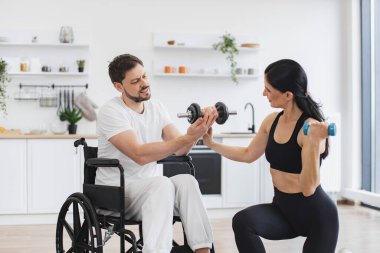 Erkek hasta, modern bir apartmanın arka planında hastalık ve yaralanma üzerine rehabilitasyona giriyor. Spor salonundaki fizyoterapist, tekerlekli sandalyedeki yetişkin bir adama evde dambıllarla nasıl egzersiz yapıldığını gösteriyor..